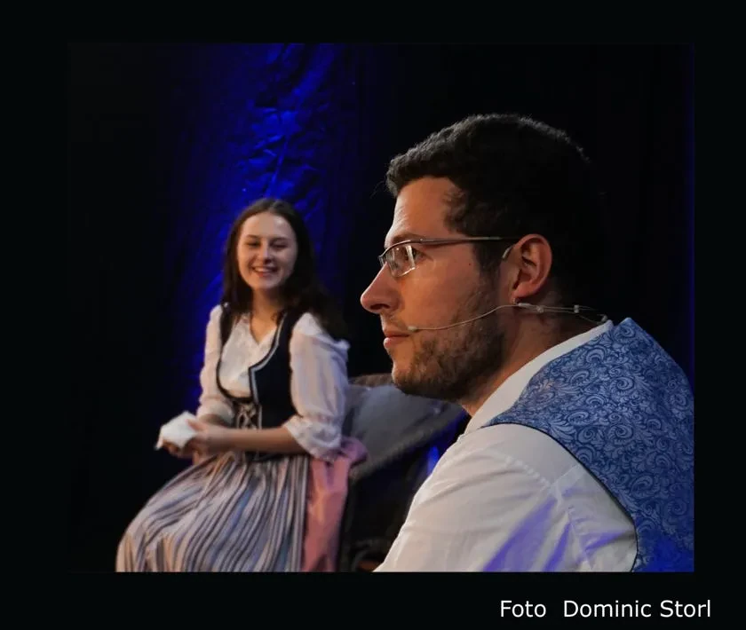 Szene auf der Bühne: vorn ein Mann mit Brille und Headset im Profil, hinter ihm lächelt eine junge Frau im Dirndl. Foto: Dominic Storl